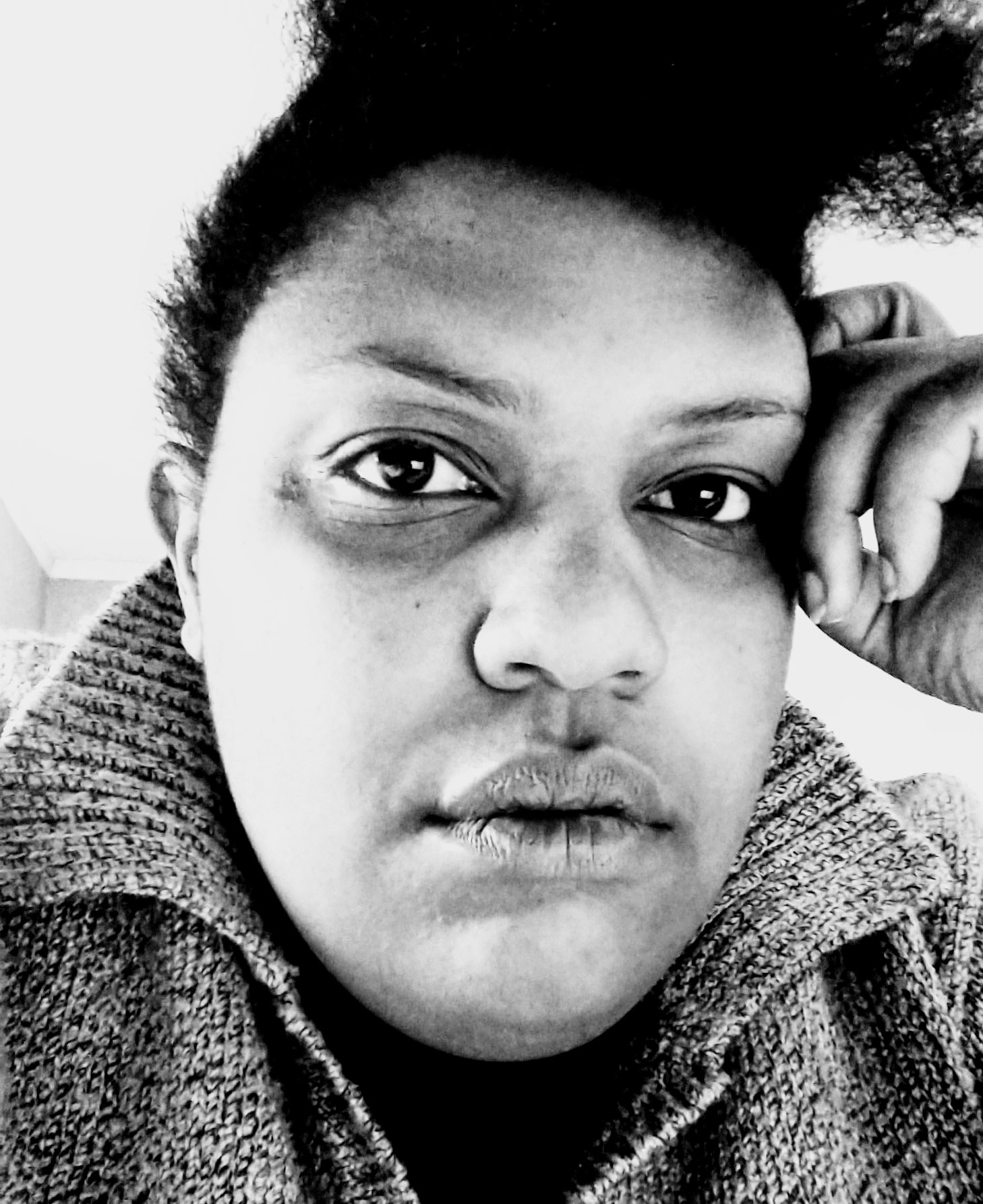 Young black woman with curly black hair cut short on the sides and long on the shop. She is wearing a tweed sweater and has a hand resting beside her eye.
