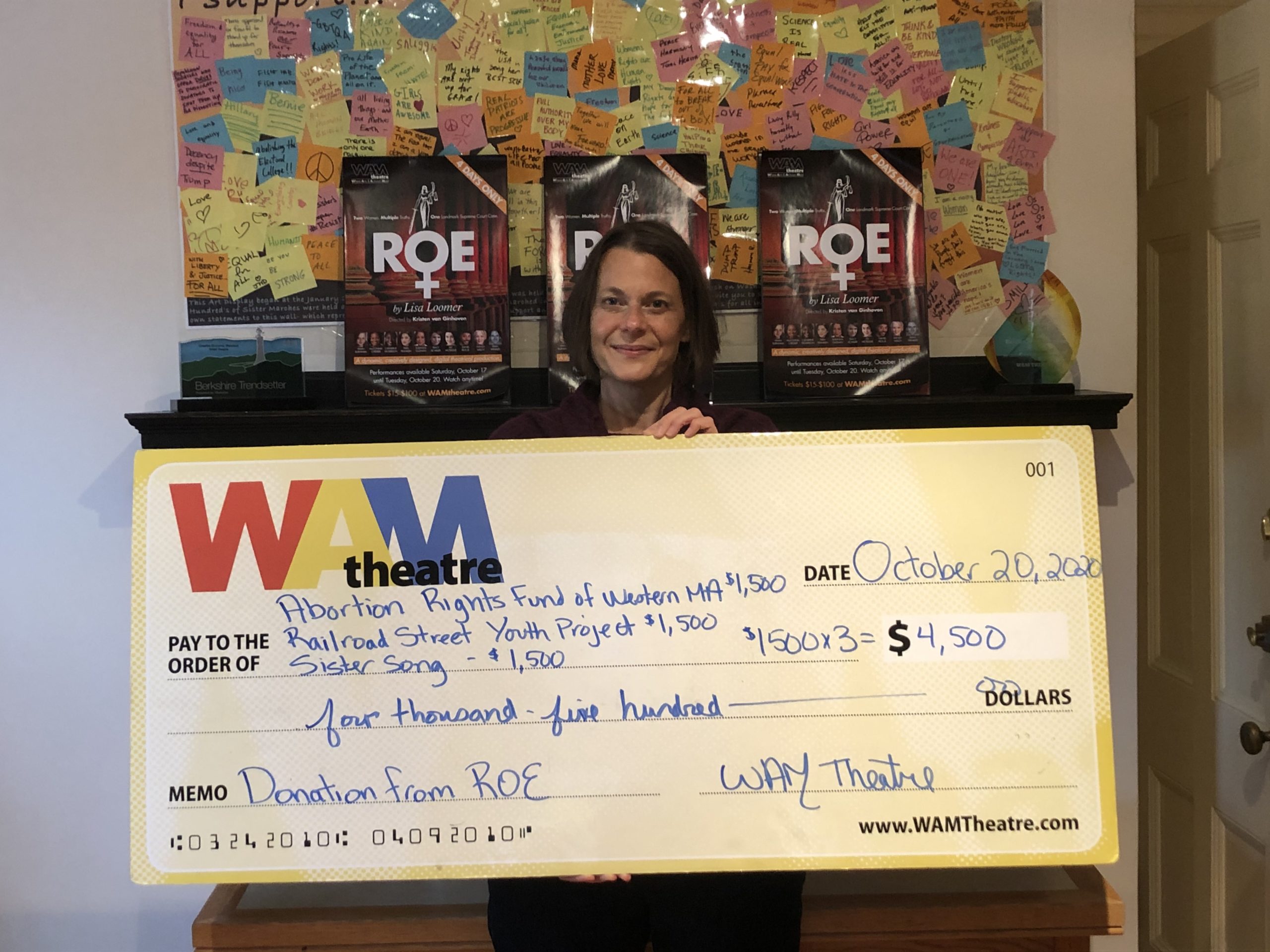 A smiling white woman with shoulder length brown hair holds a giant check representing the donations WAM made to its 2020 beneficiaries.