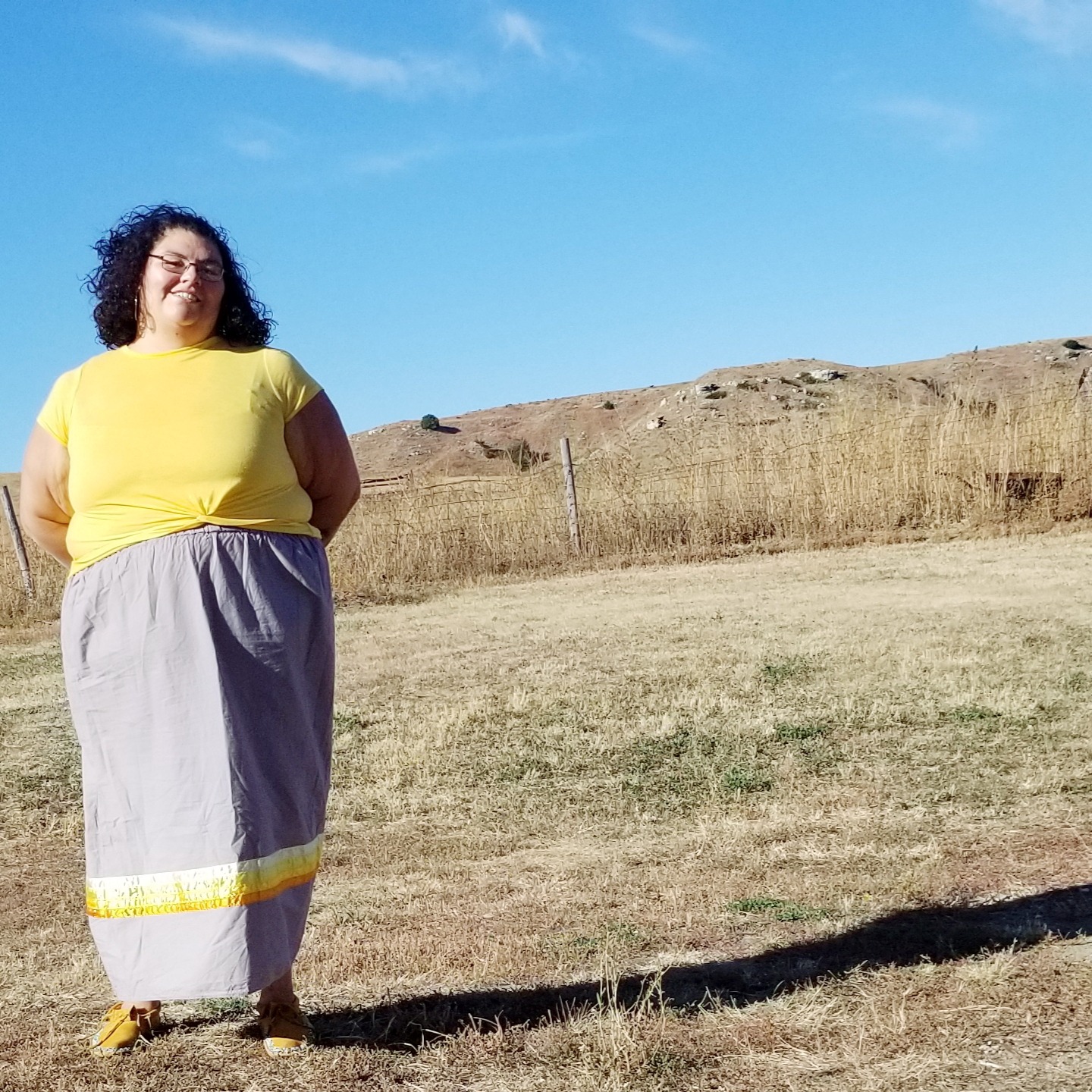 miling young indigenous woman with curly shoulder length brown hair. She is wearing eyeglasses and a light yellow top and a long gray skirt with bands of white and yellow towards the bottom.