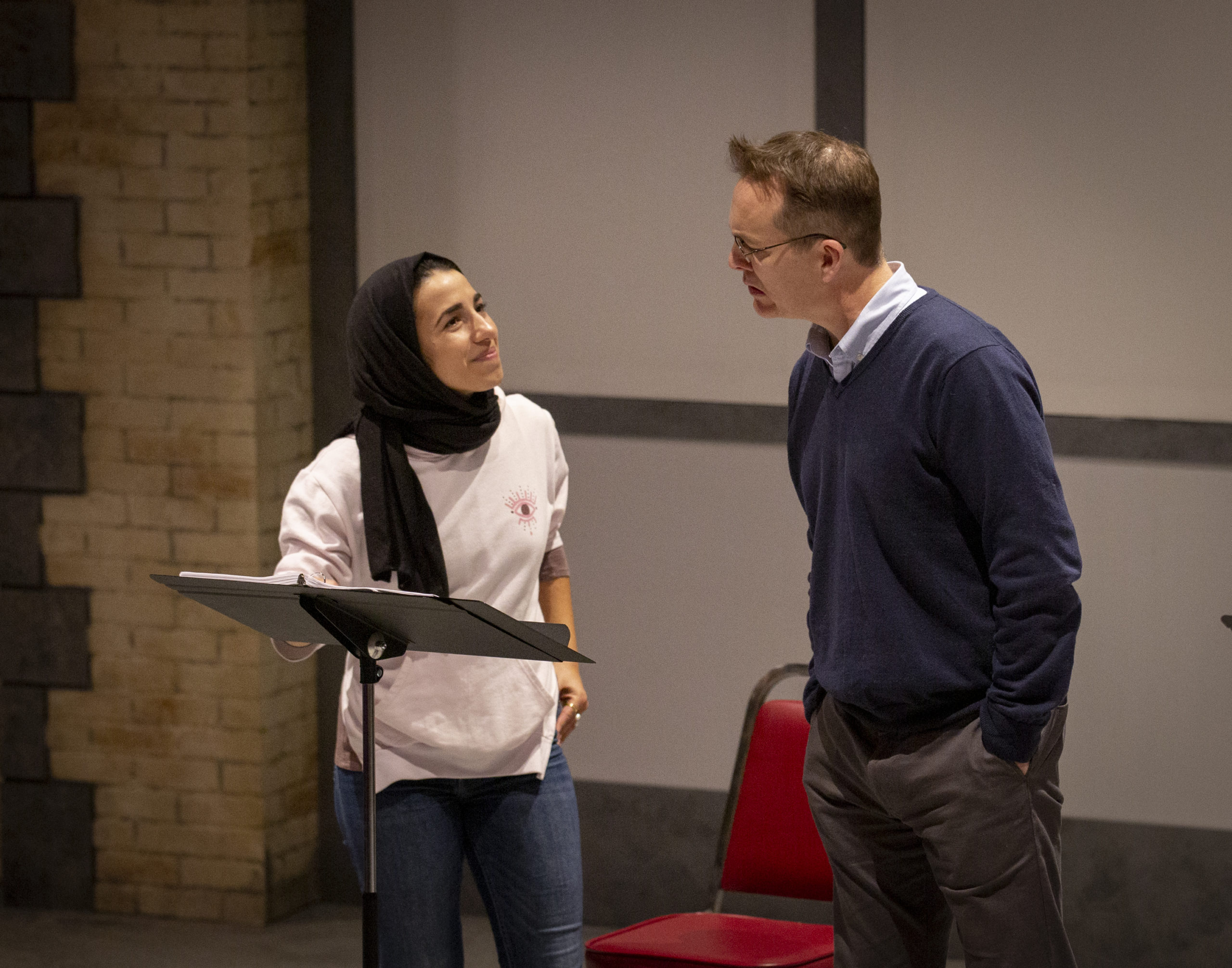Young Arab woman wearing a black headscarf, white sweatshirt, and jeans on the left; in conversation with a taller white man wearing glasses, a navy v-neck sweater over a white collared shirt and grey slacks.