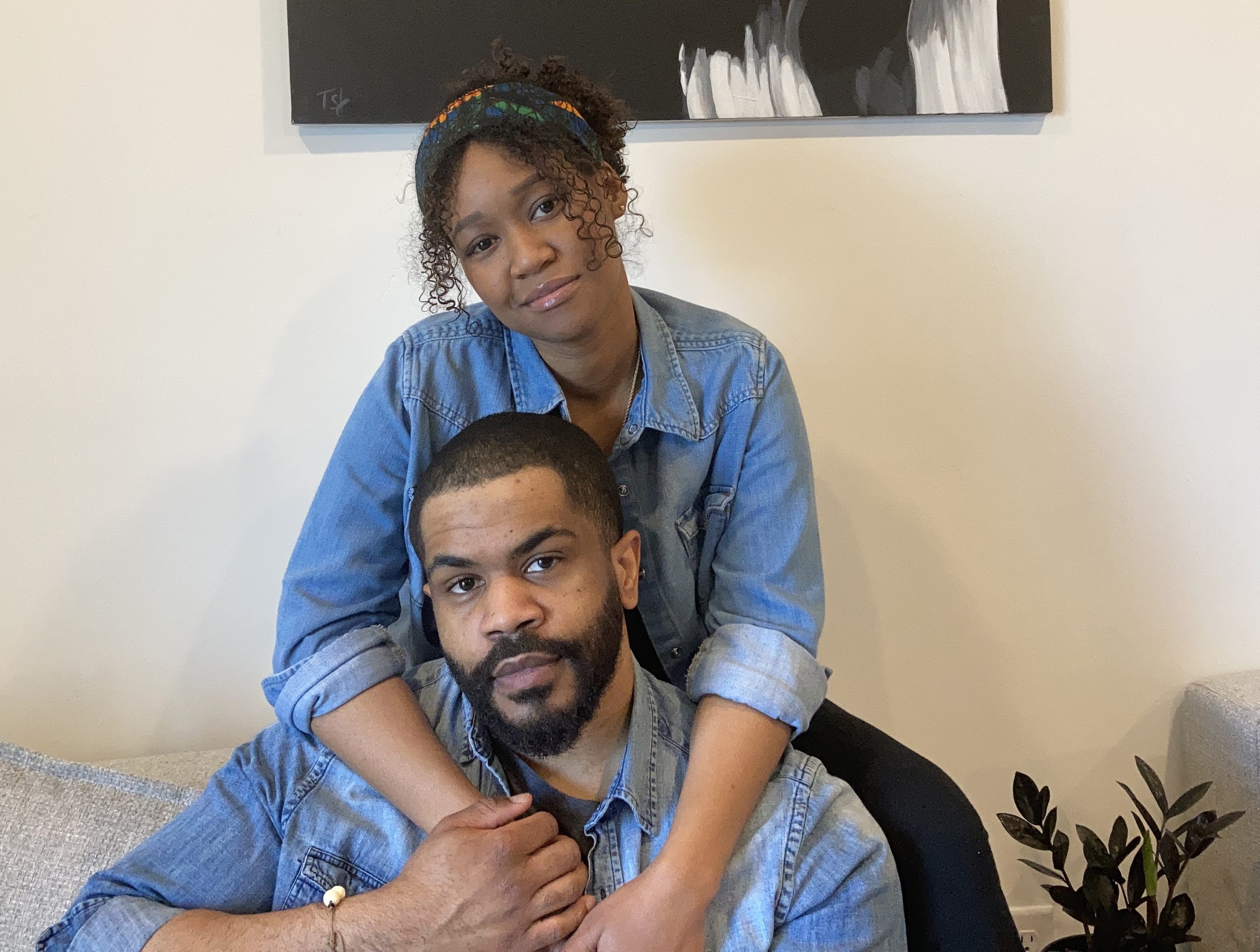 A heterosexual Black couple seated in front of a white wall. Both are wearing washed denim work shirts. She is behind him with her arms around his neck. He has a close-cropped beard and mustache.